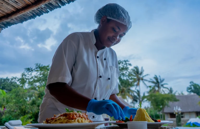 Ikumbi chef plating a dish outdoors under the thatched eave