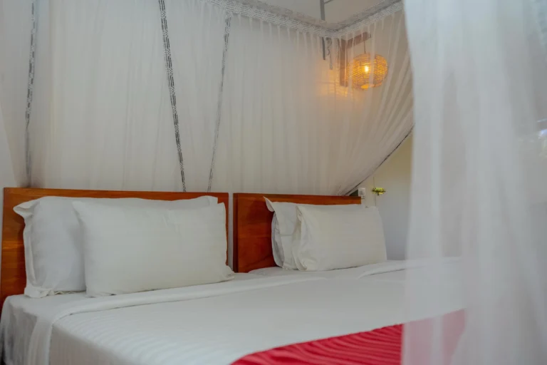 Two beds with white linen under a canopy mosquito net, lit by a woven wall lamp